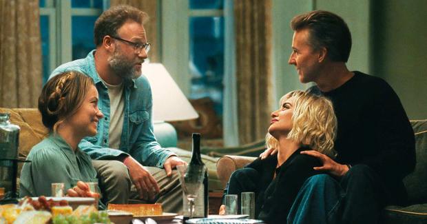 Seth Rogen, Edward Norton, Shana Hiatt, Olivia Wilde: Cena do filme 'Ghosted' com quatro pessoas conversando em uma sala de estar ao redor de uma mesa com petiscos e vinho.