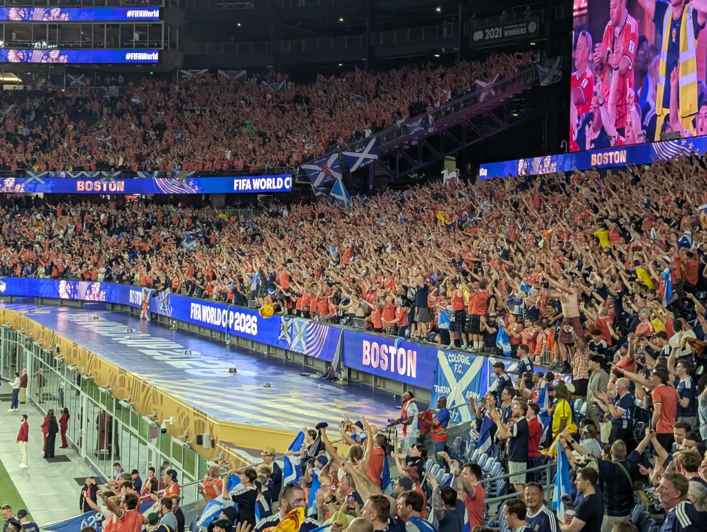 Uma grande multidão de torcedores com camisas laranjas e azuis comemorando em um estádio de futebol com painéis eletrônicos exibindo 'BOSTON' e 'FIFA WORLD CUP 2026'.