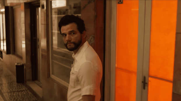 Wagner Moura: Cena de um filme mostrando um homem barbudo com camisa branca olhando para trás em um corredor com iluminação alaranjada.