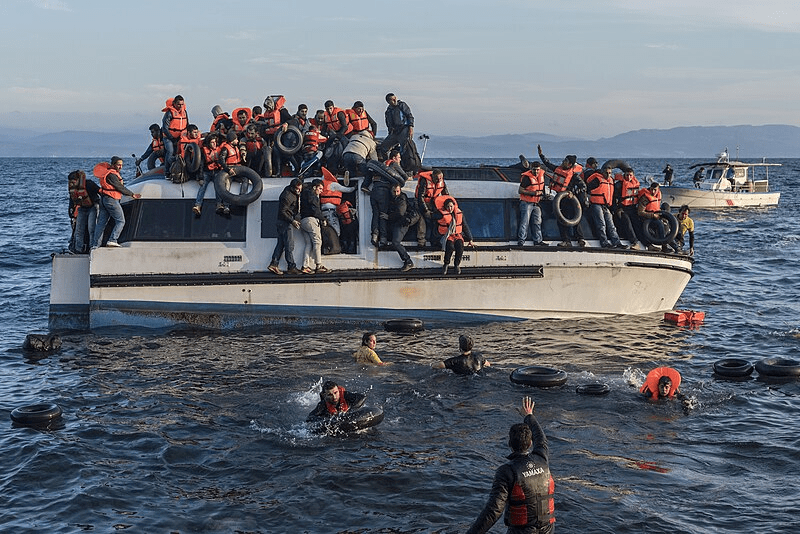 Uma balsa superlotada com refugiados e migrantes usando coletes salva-vidas no mar Mediterrâneo, com algumas pessoas na água tentando alcançar a embarcação ou usando boias.