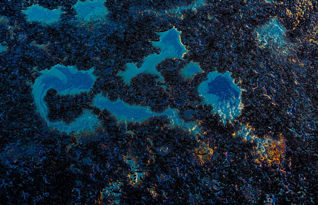 Fotografia em close de manchas de óleo ou combustível sobre uma superfície de asfalto molhada, criando padrões azuis e iridescentes.