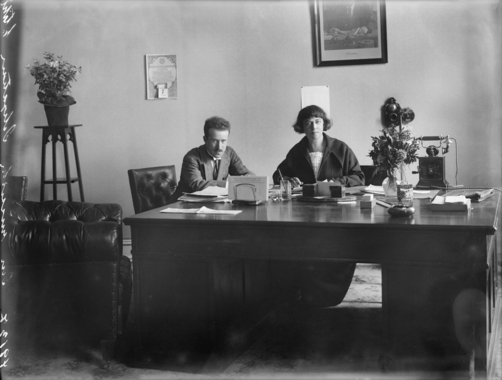 António Ferro e Fernanda de Castro: Uma fotografia antiga em preto e branco de um homem e uma mulher sentados atrás de uma grande mesa de escritório de madeira, cercados por documentos e um telefone antigo.