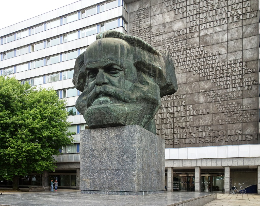 Karl Marx: Uma fotografia do Monumento a Karl Marx, uma escultura gigante de bronze da cabeça de Marx em Chemnitz, Alemanha, com uma parede ao fundo exibindo a frase 'Proletários de todos os países, uni-vos!' em vários idiomas.