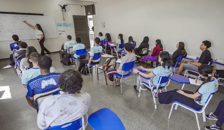 desconhecido: Uma professora escrevendo no quadro branco enquanto alunos assistem à aula em uma sala de aula.