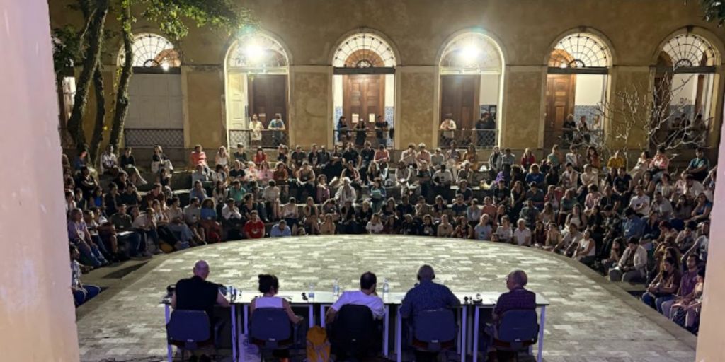 Uma assembleia ou debate público ocorrendo à noite em um pátio circular com uma grande plateia sentada em degraus ao fundo e uma mesa de palestrantes em primeiro plano.