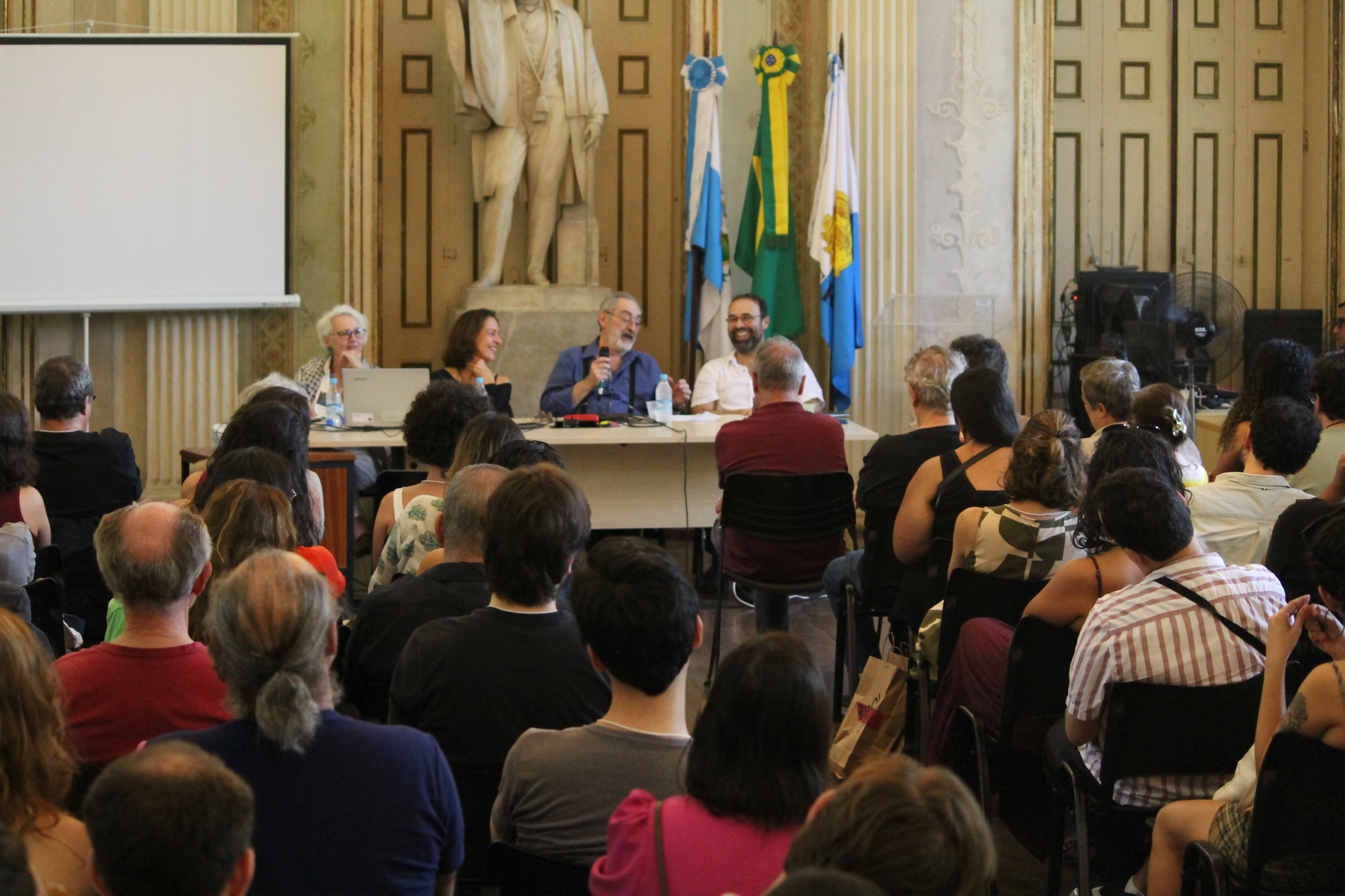 desconhecido: Uma conferência ou debate ocorrendo em um salão histórico com uma plateia sentada e quatro palestrantes à mesa.