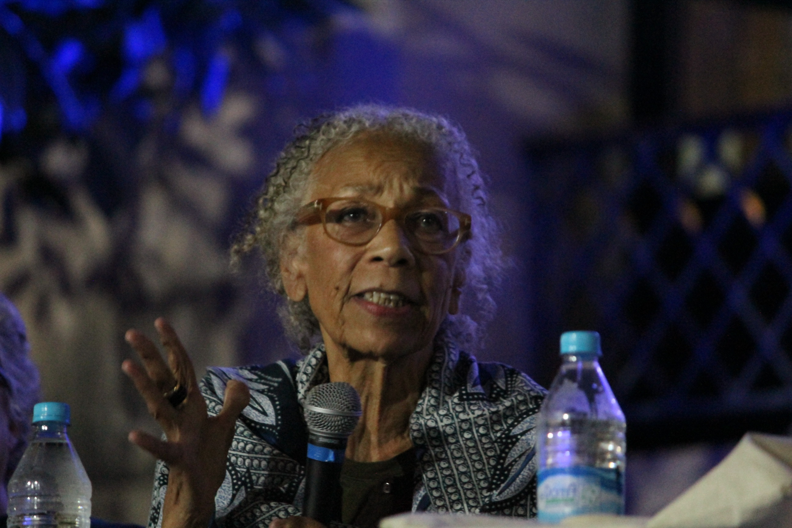 Maria Conceição Evaristo de Brito: Uma mulher negra de cabelos grisalhos e óculos falando ao microfone em um evento noturno.