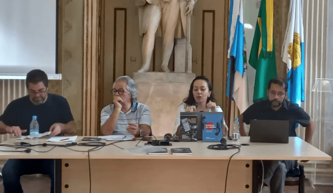 unknown: Quatro pessoas sentadas à mesa em um debate ou seminário, com livros sobre a mesa e bandeiras ao fundo.