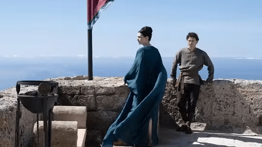 Rachel Weisz, Tom Hiddleston: Cena de filme ou série de época mostrando uma mulher com vestido longo azul e um homem em trajes rústicos em um terraço de pedra com vista para o mar.