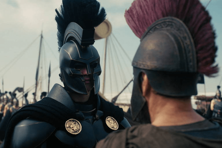 Dois guerreiros usando capacetes gregos clássicos com cristas de penas se encarando em um cenário de navios antigos.