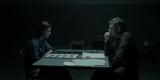 Maria de Medeiros, Marcello Antony: Uma cena de filme em estilo noir mostrando duas pessoas sentadas em uma mesa em uma sala de interrogatório escura.