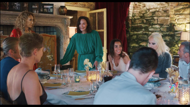 Kristin Scott Thomas: Uma cena de filme ou série mostrando um grupo de pessoas reunidas em torno de uma mesa de jantar em um ambiente rústico e sofisticado.