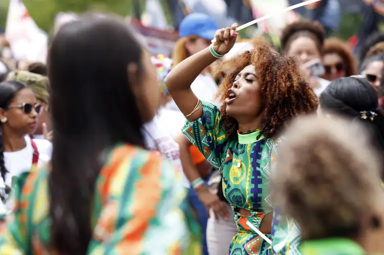 Uma mulher negra com cabelos cacheados e roupas coloridas participa de um evento ou manifestação ao ar livre, segurando um bastão e gritando.