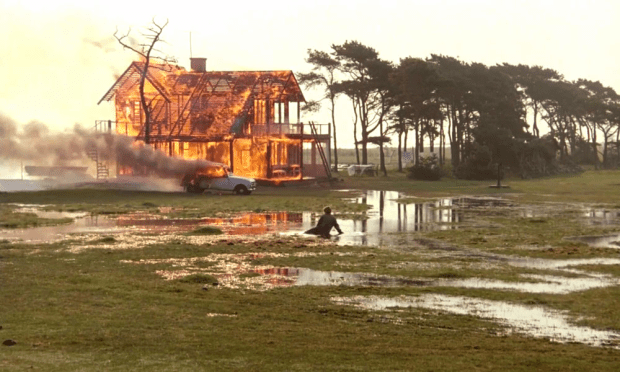 Erland Josephson: Uma casa de madeira em chamas em um campo aberto com áreas alagadas, cena do filme 'O Sacrifício' de Andrei Tarkovsky.