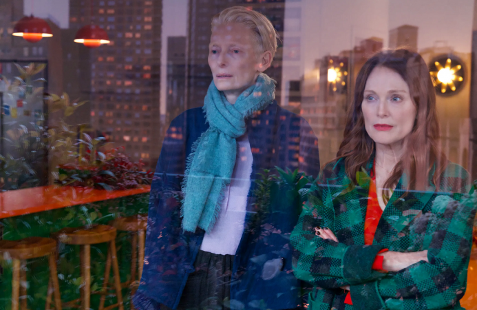 Tilda Swinton, Julianne Moore: Duas mulheres em pé dentro de um ambiente com reflexos de janelas e luzes de uma cidade ao fundo. Trata-se de uma cena do filme 'O Quarto ao Lado' (The Room Next Door).