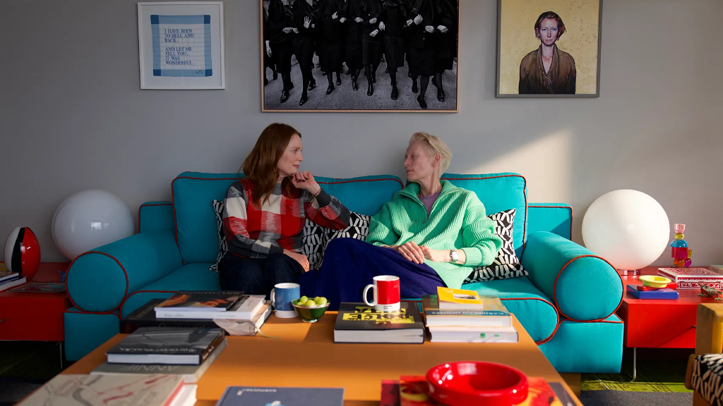 Julianne Moore, Tilda Swinton: Uma cena de filme onde duas mulheres conversam sentadas em um sofá colorido com vários livros sobre a mesa à frente.