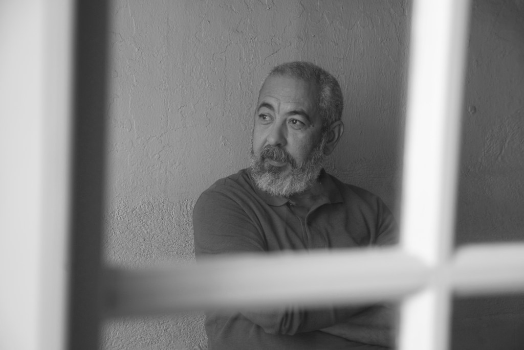 Leonardo Padura: Retrato em preto e branco de um homem com barba grisalha, visto através de uma estrutura que se assemelha a uma moldura de janela.