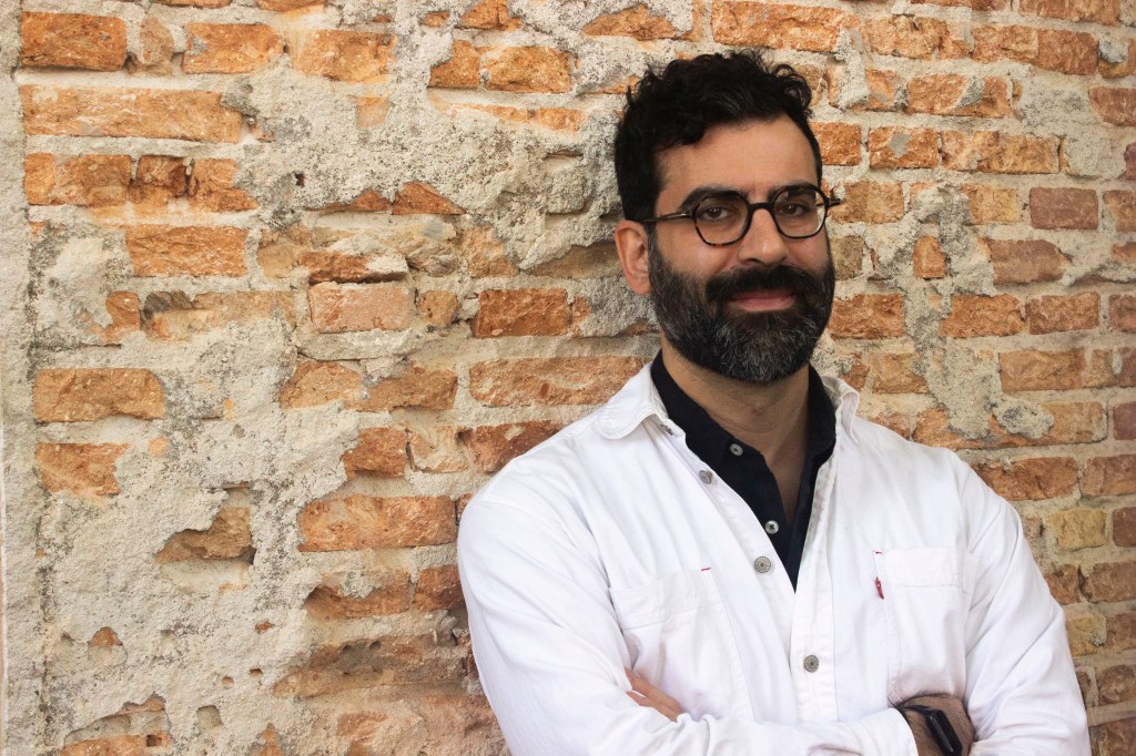 Valter Hugo Mãe: Homem de óculos, barba e camisa branca com braços cruzados posando em frente a uma parede de tijolos.