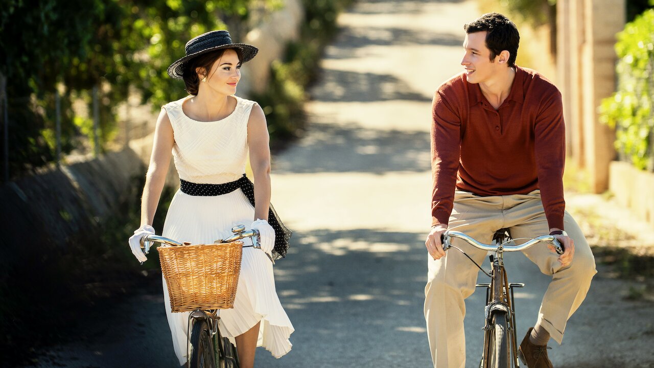 Shailene Woodley, Callum Turner: Uma mulher vestida de branco com chapéu e um homem vestindo blusa vermelha andam de bicicleta lado a lado em uma estrada ensolarada.