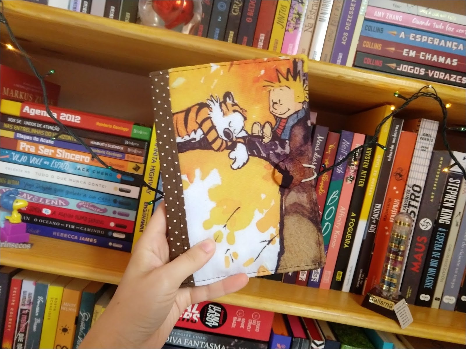 Mão segurando uma capa de tecido artesanal com estampa de Calvin e Haroldo em frente a uma estante cheia de livros.