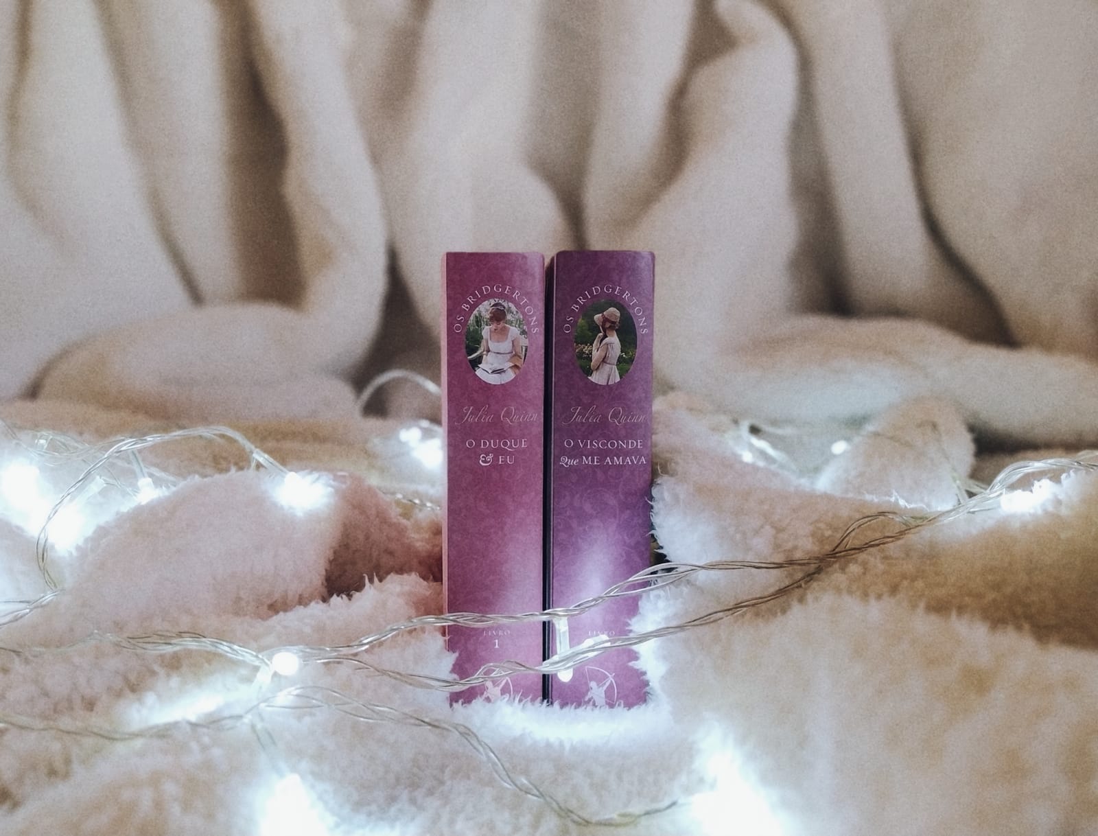 Lombadas dos livros 'O Duque e Eu' e 'O Visconde que me Amava' da série Os Bridgertons, posicionadas sobre uma manta felpuda com luzes decorativas (fairy lights).