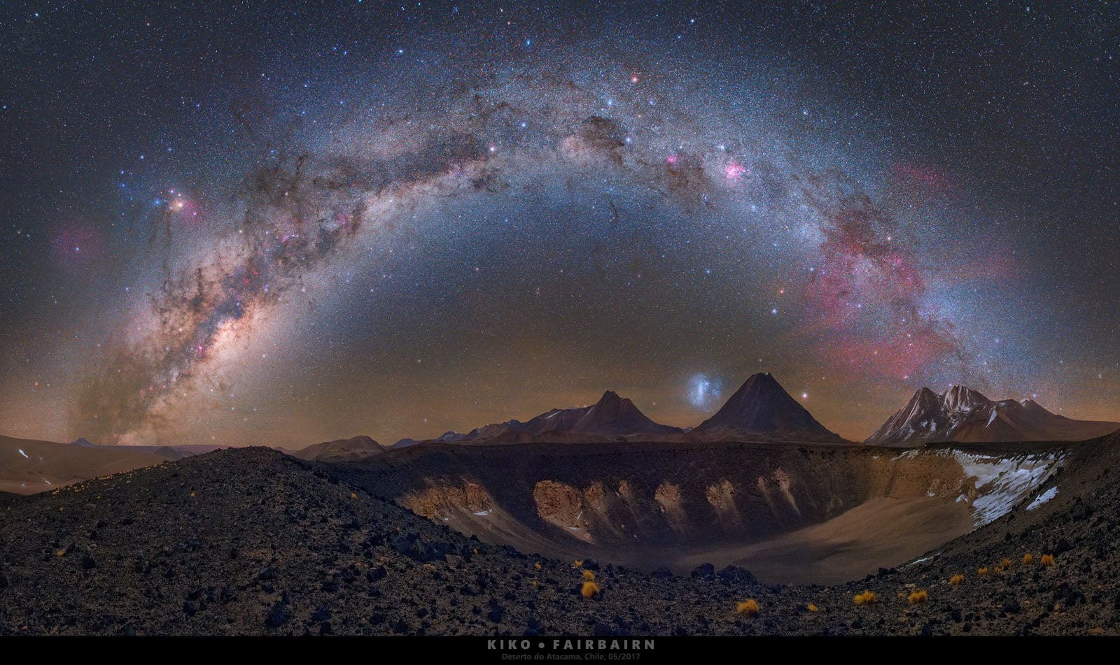 A panoramic night view of the Milky Way arching over a volcanic crater and mountain range in the Atacama Desert, Chile.