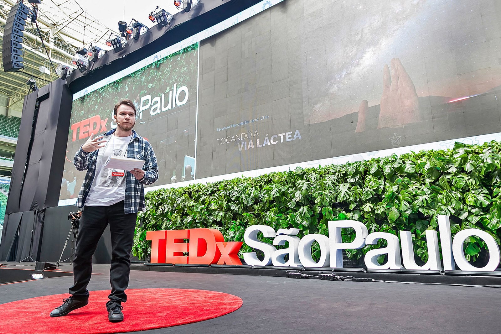 A man in a plaid shirt and white t-shirt stands on a red circular rug on a stage, gesturing while holding notes in front of a large screen displaying "TEDx São Paulo" and an image of the Mano del Desierto sculpture.
