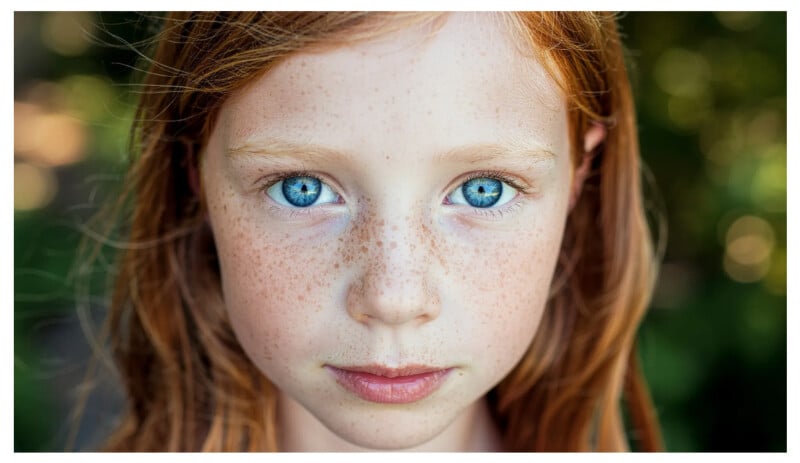 Close-up portrait of a young girl with red hair, bright blue eyes, and freckles, looking directly at the camera with a serious expression. The background is softly blurred with greenery.