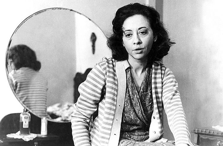 Fernanda Montenegro: Fotografia em preto e branco de uma mulher sentada em frente a um espelho circular, vestindo um cardigã listrado.