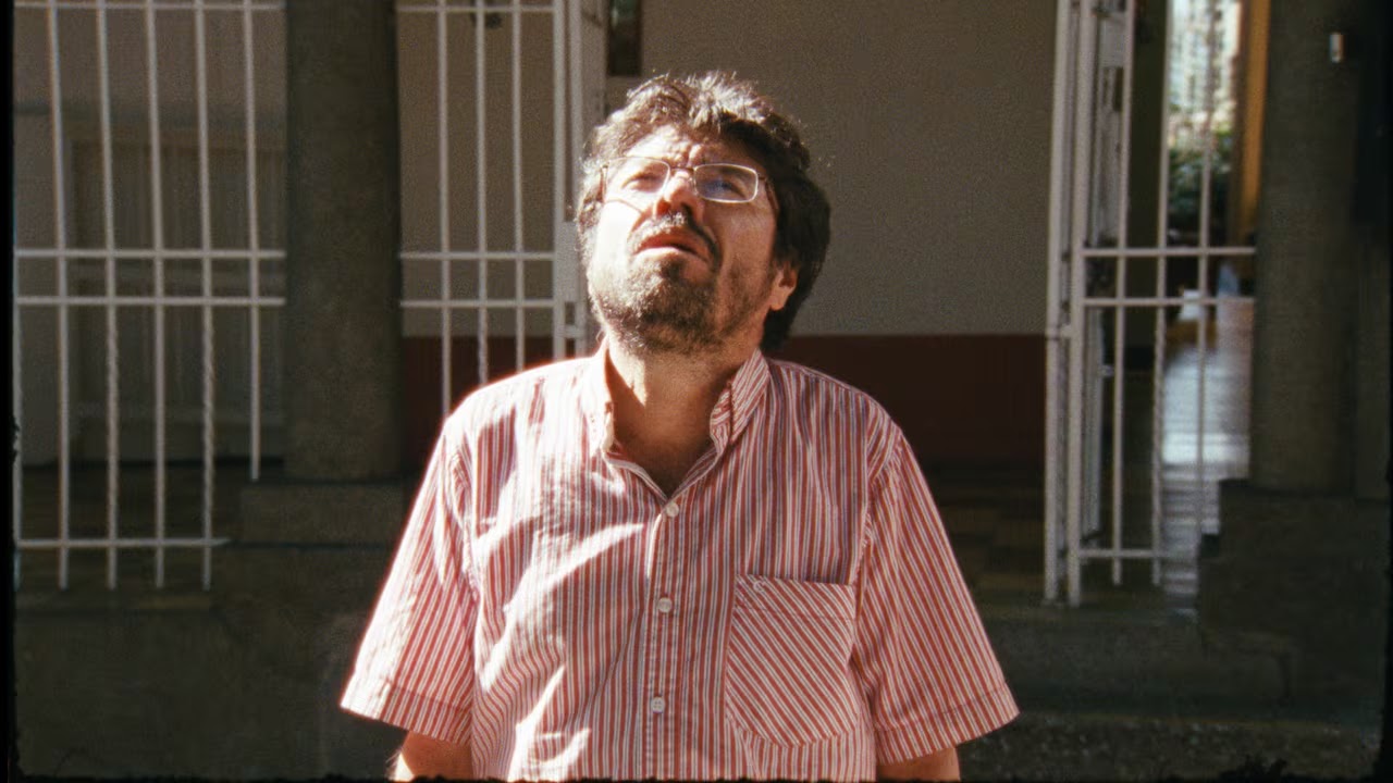 Ettore Scola: Homem de óculos e camisa listrada olhando para cima em um ambiente externo com grades ao fundo.