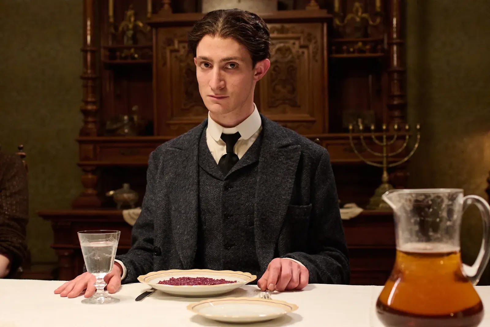 Joel Basman: Uma fotografia de cena do filme 'Kafka', mostrando o ator Joel Basman como Franz Kafka sentado à mesa de jantar.