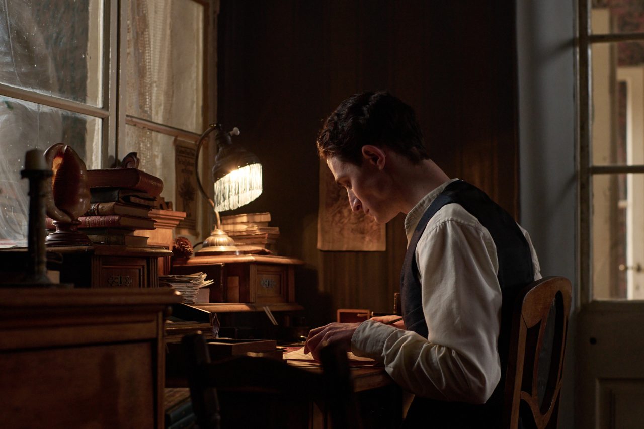 Franz Kafka (portrayed by Sabin Tambrea): A man in period clothing sitting at a rustic wooden desk, writing by the light of a small lamp in a dimly lit room.