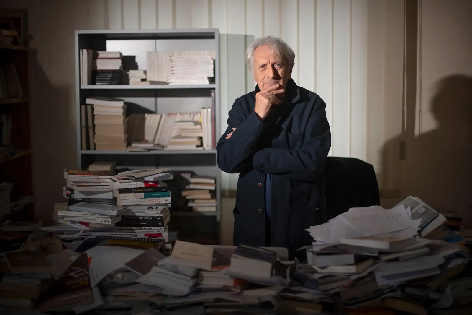 António Lobo Antunes: Retrato de um homem idoso com a mão no queixo, cercado por pilhas de livros e papéis em um escritório.