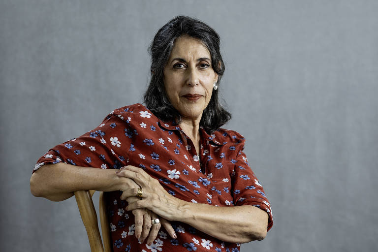 Lúcia Hiratsuka: Retrato de uma mulher de meia-idade vestindo uma blusa floral vermelha, sentada em uma cadeira contra um fundo cinza.