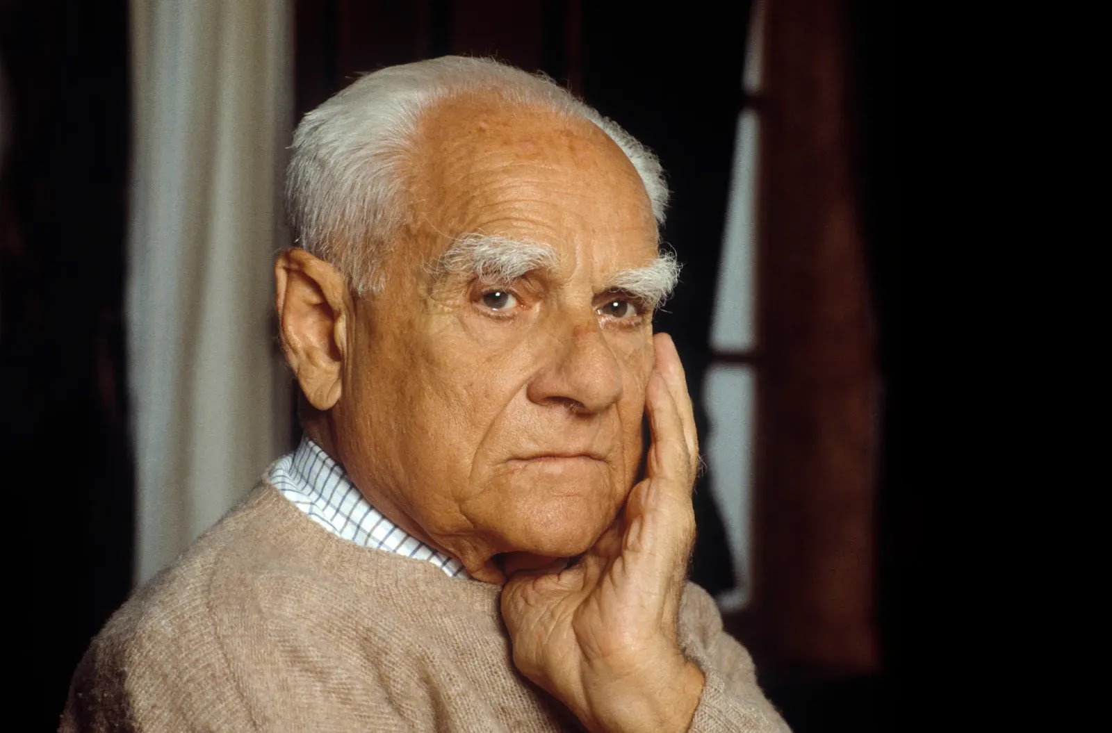 Alberto Moravia: Retrato de um homem idoso com sobrancelhas brancas proeminentes apoiando o rosto na mão.