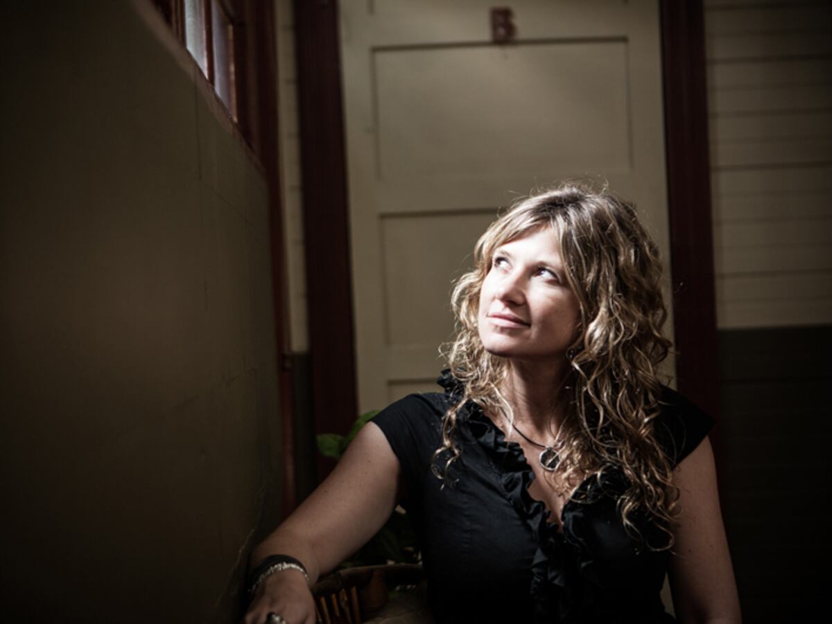 unknown: Uma mulher loira de cabelos cacheados olhando para cima, em um ambiente interno com iluminação dramática vinda da esquerda.