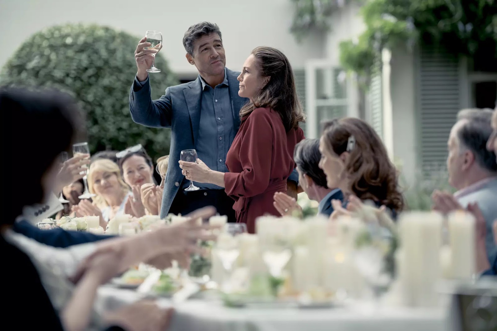 Kyle Chandler, Diane Lane: Um homem faz um brinde com uma taça de vinho ao lado de uma mulher em um jantar ao ar livre com convidados.