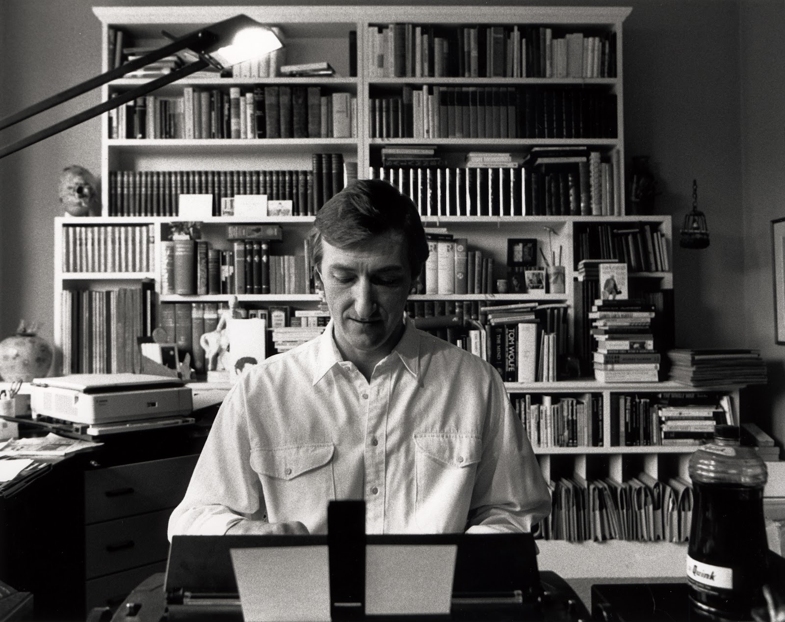 Julian Barnes: Fotografia em preto e branco de um homem sentado em frente a uma máquina de escrever, com uma grande estante de livros ao fundo.