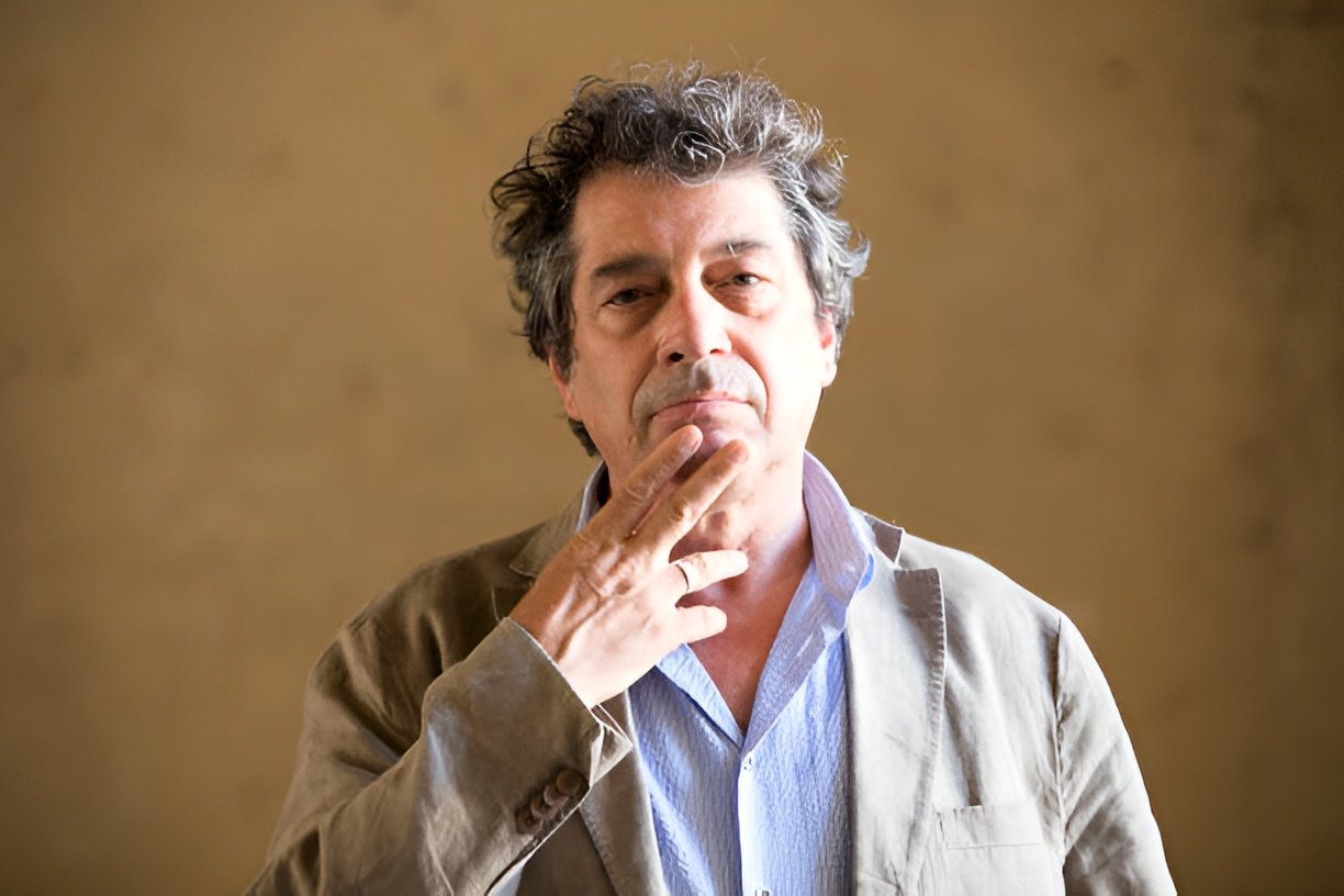 António Pedro Vasconcelos: Retrato de um homem de meia-idade com cabelos grisalhos e cacheados, vestindo um paletó bege sobre uma camisa azul, com a mão no queixo em pose reflexiva.