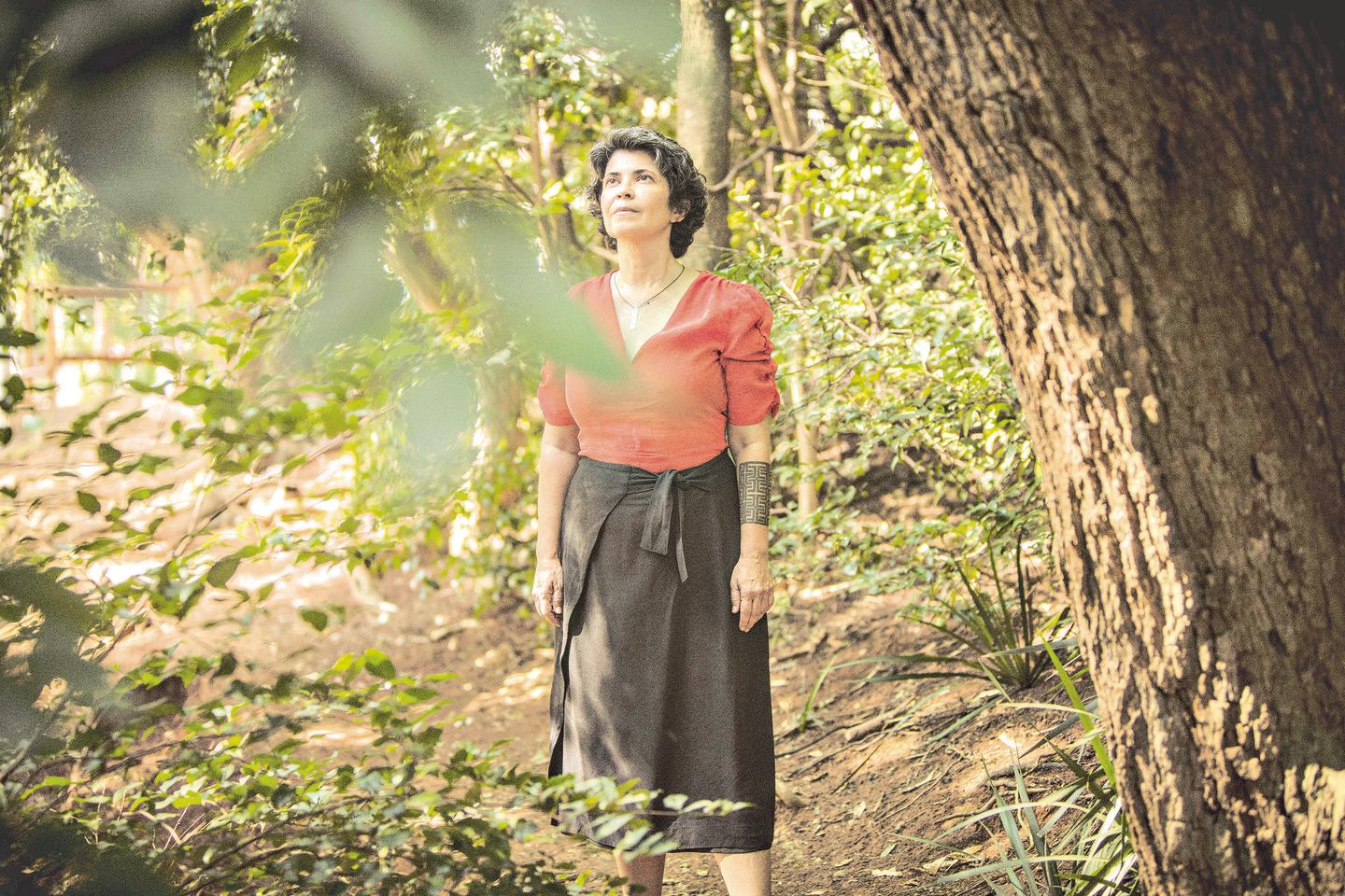 unknown: Uma mulher de cabelos curtos grisalhos, vestindo blusa vermelha e saia preta, em pé em um ambiente arborizado, olhando para cima.